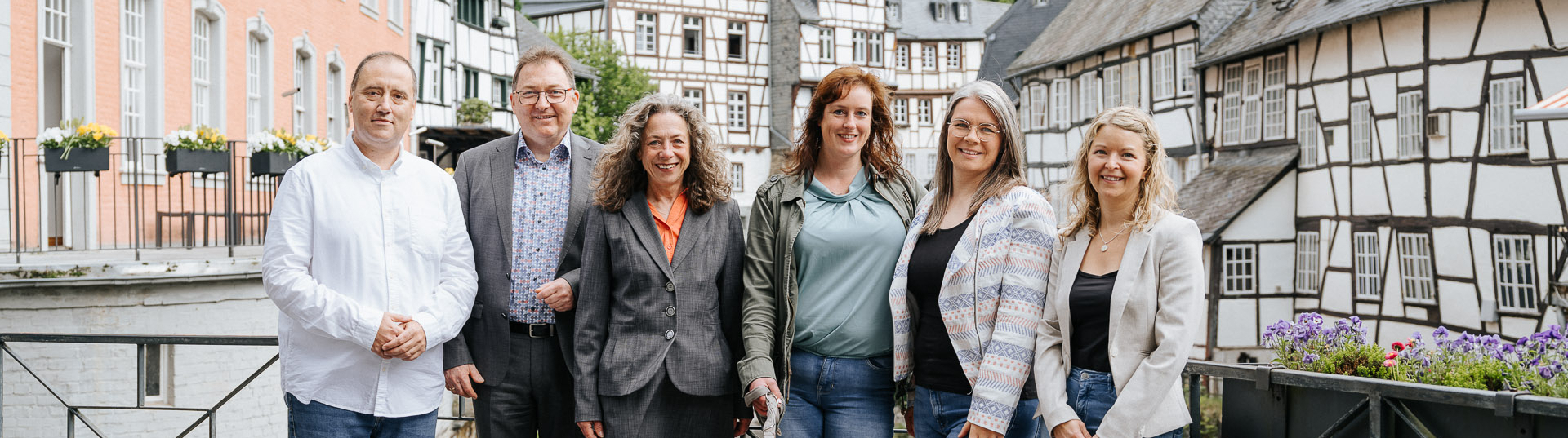 Gruppe von Mitarbeitenden von immoGrafik steht gemeinsam mit zwei Hunden auf einer Brücke vor historischen Fachwerkhäusern in einer Altstadt.