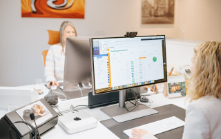 Zwei Mitarbeiterinnen arbeiten an einem Schreibtisch im Büro von immoGrafik. Auf einem großen Monitor ist ein Kundensupport-Dashboard geöffnet, während im Vordergrund eine Person mit Tastatur arbeitet und im Hintergrund eine Kollegin am Computer sitzt.