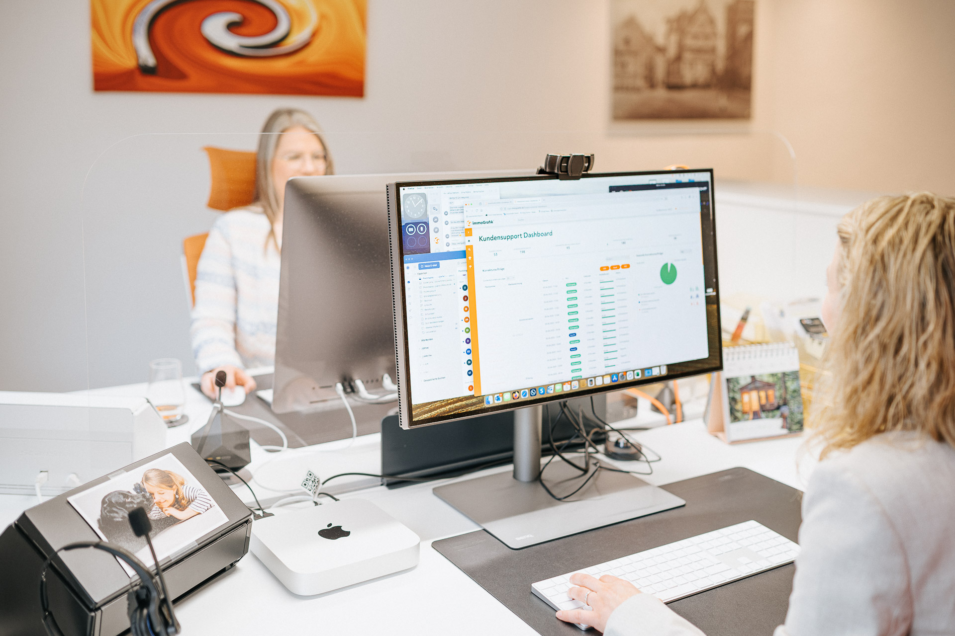 Zwei Mitarbeiterinnen arbeiten an einem Schreibtisch im Büro von immoGrafik. Auf einem großen Monitor ist ein Kundensupport-Dashboard geöffnet, während im Vordergrund eine Person mit Tastatur arbeitet und im Hintergrund eine Kollegin am Computer sitzt.