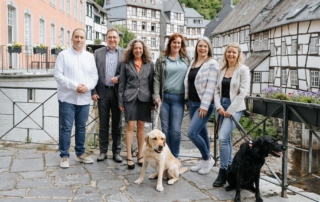 Gruppe von Mitarbeitenden von immoGrafik steht gemeinsam mit zwei Hunden auf einer Brücke vor historischen Fachwerkhäusern in einer Altstadt.