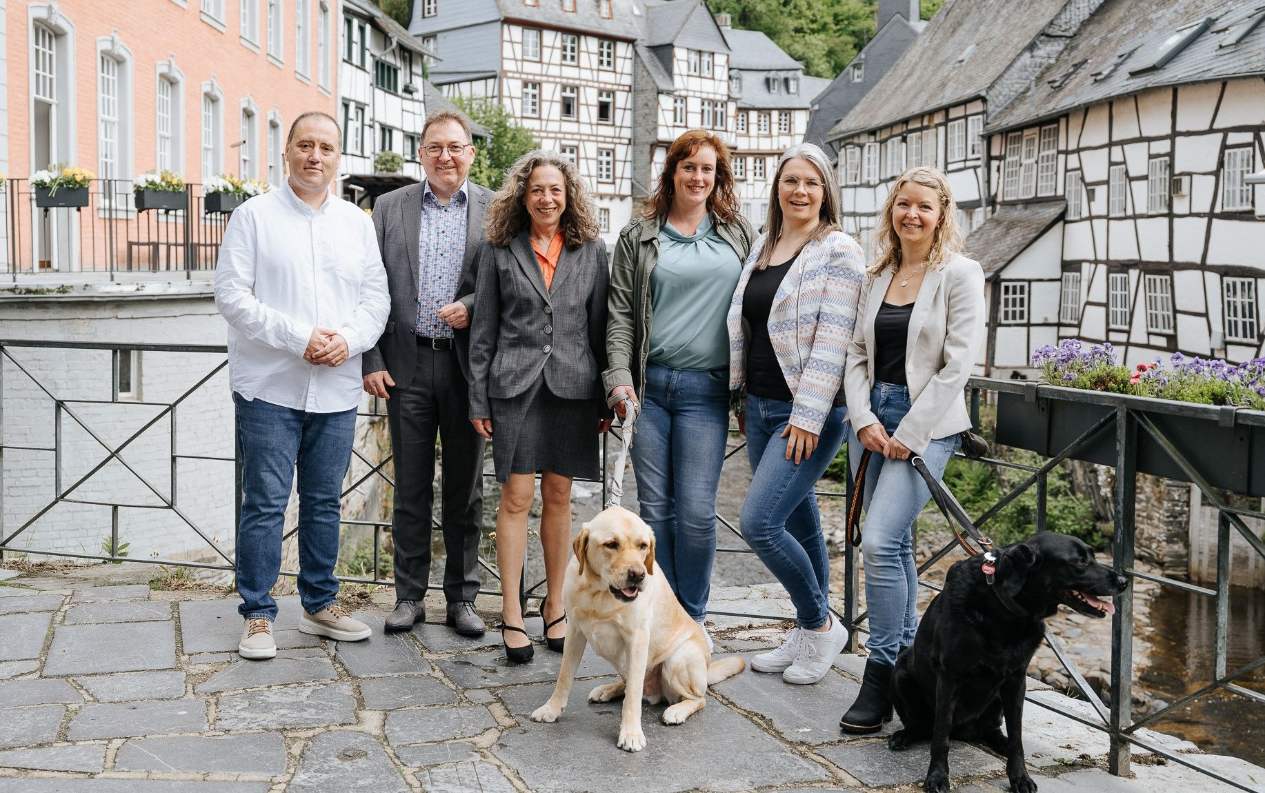 Gruppe von Mitarbeitenden von immoGrafik steht gemeinsam mit zwei Hunden auf einer Brücke vor historischen Fachwerkhäusern in einer Altstadt.
